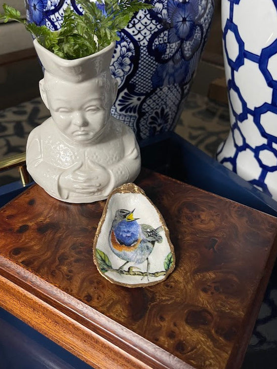 Decorative oyster shell with a single blue bird on a wooden surface, next to a bust-shaped vase and another decorative item.