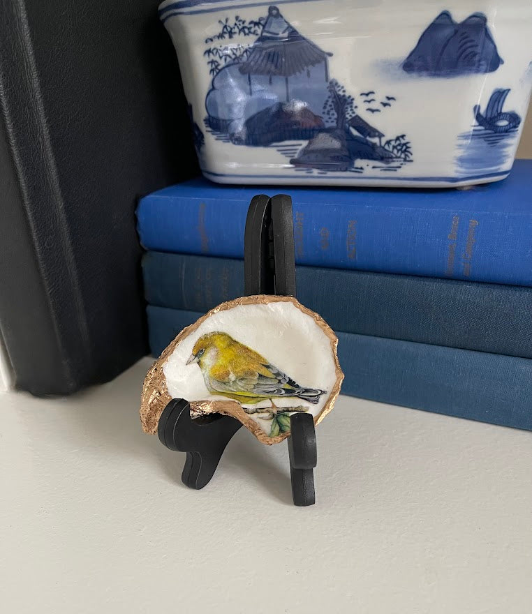 Decorative oyster shell with a yellow bird on a stand, with a blue and white ceramic piece in the background.