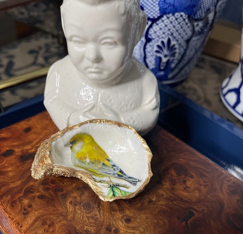 Decorative shell with a yellow bird on a wooden surface, with a white ceramic bust in the background.